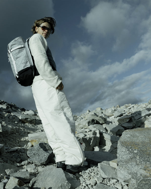 Woman wearing the Db Backcountry Backpack 25L in White Noise while standing on rocky mountain terrain.