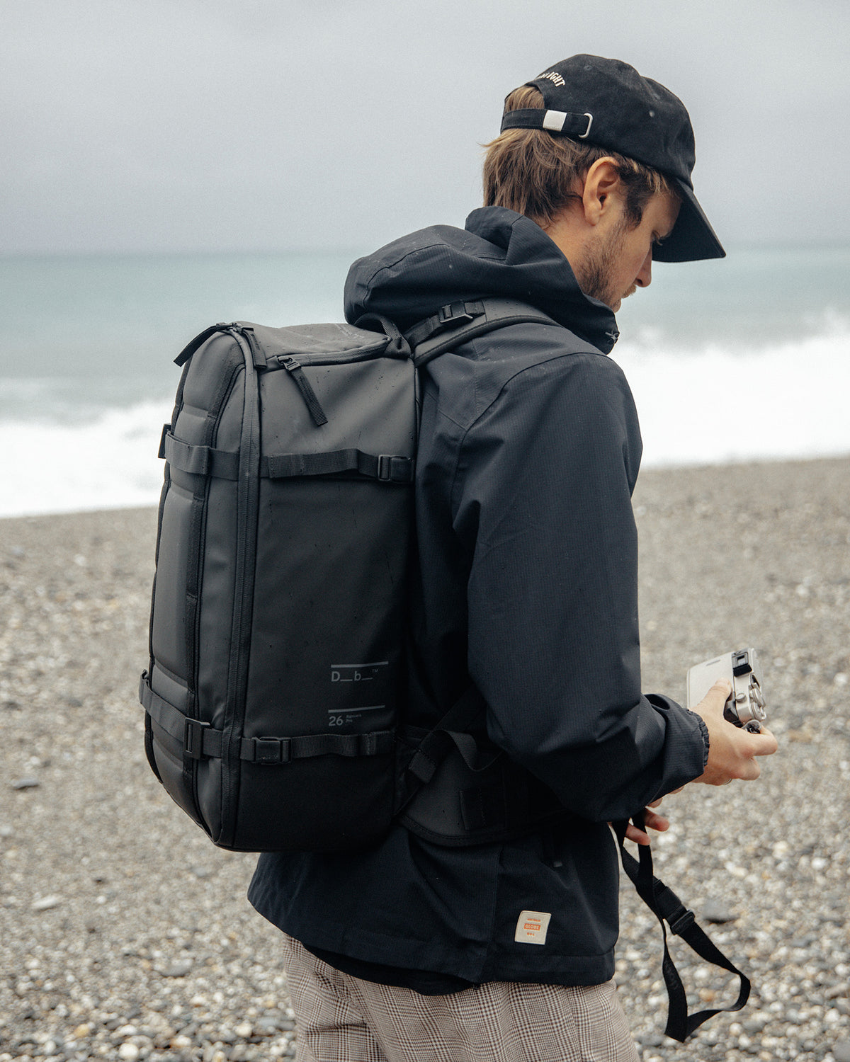 Svart Db Ramverk Pro Ryggsekk 26L båret av en fotograf som holder et kamera på en steinstrand.